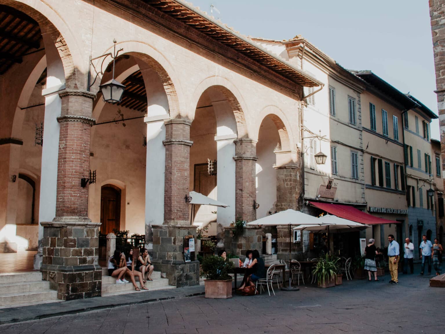 Geheimtipp für Toskana-Reisende: Das kleine Dörfchen Montalcino - hier Arcaden, unter denen ein paar Jugendliche sitzen, daneben ein gemütliches Café mit Sonnenschirmen auf der Außenterrasse.