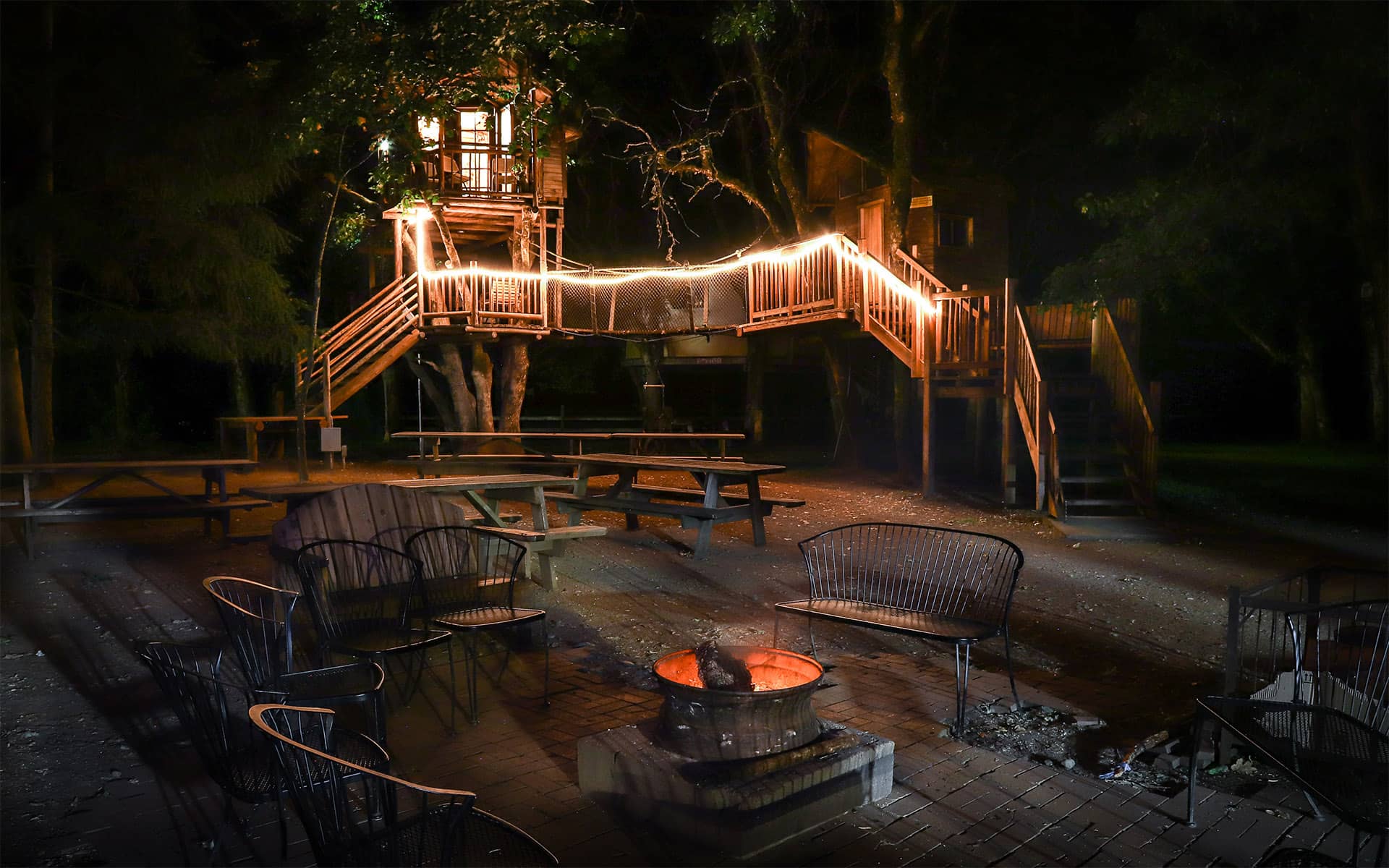 Ein Baumhaus mit Holzsteg und Lichterkette im Hintergrund einer Sitzlounge im freien mit Lagerfeuer.